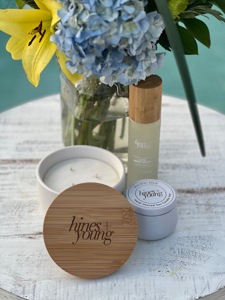 ceramic candle, capri honeymoon linen spray, and tin candle on table in front of blue and yellow flowers